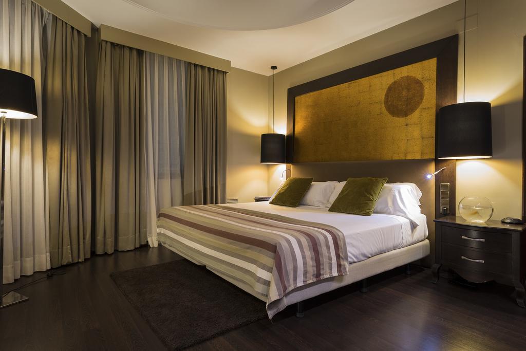 Only You Hotel Valencia Luaran gambar The photo shows a modern bedroom that features a large bed with two green accent pillows and a striped bedspread. The room has a warm, neutral color scheme, highlighted by soft lighting from two bedside lamps. The wall behind the bed has an artistic 